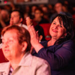 Smiling woman in a dark blue outfit claps while seated in a crowded theater with red seats behind her.