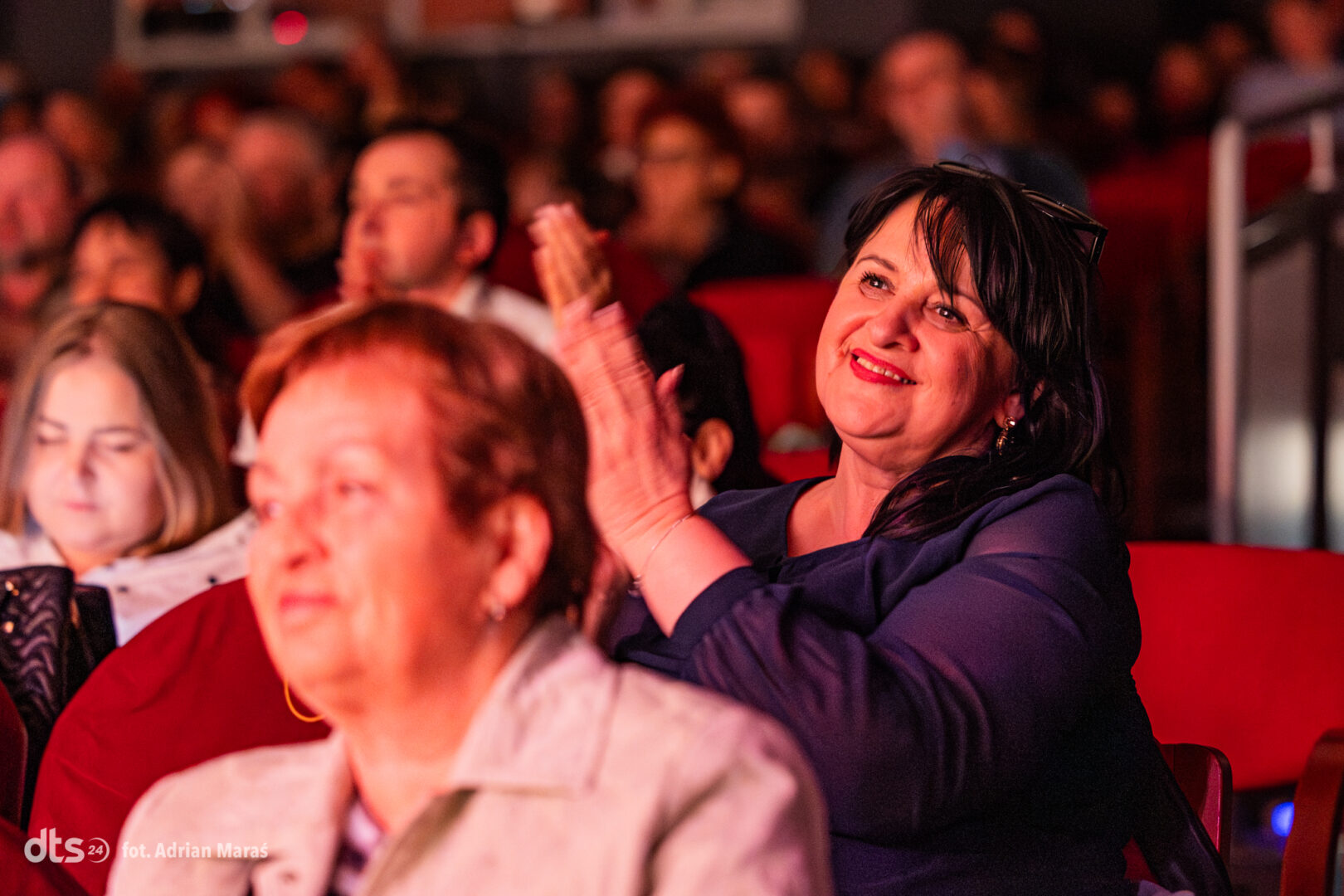 Smiling woman in a dark blue outfit claps while seated in a crowded theater with red seats behind her.