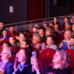 Audience in a dimly lit theater with red seats, clapping and watching a performance.
