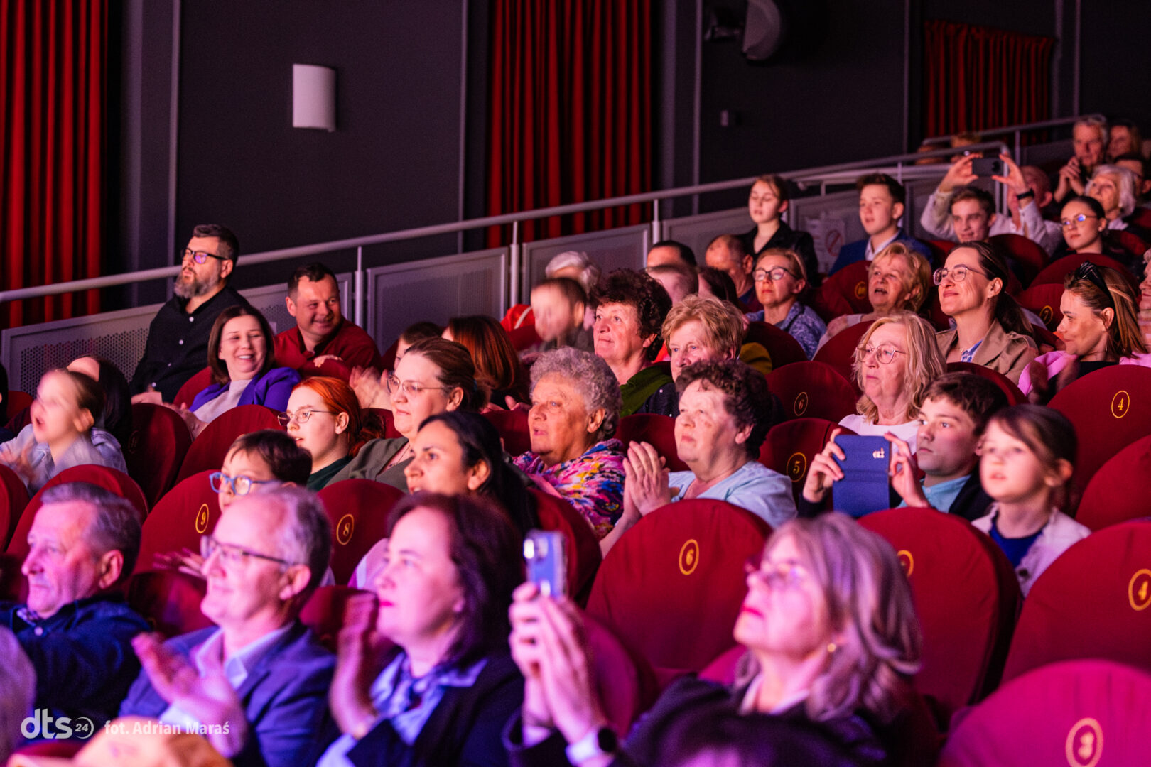 Audience in a dimly lit theater with red seats, clapping and watching a performance.