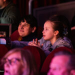 Two kids sit in red theater seats, one holding a phone to film the show while the other watches intently.
