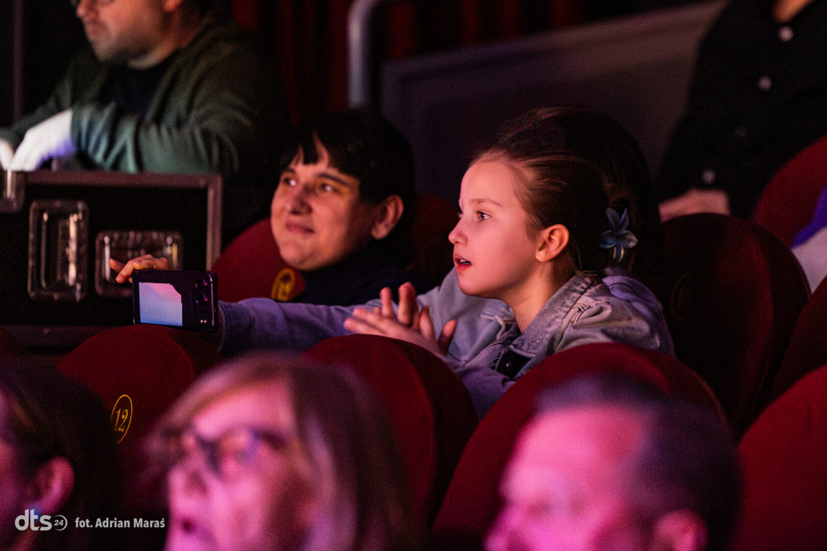 Two kids sit in red theater seats, one holding a phone to film the show while the other watches intently.