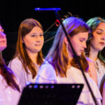 Group of young women in white shirts standing on a dimly lit stage with purple lighting, singing in a choir.