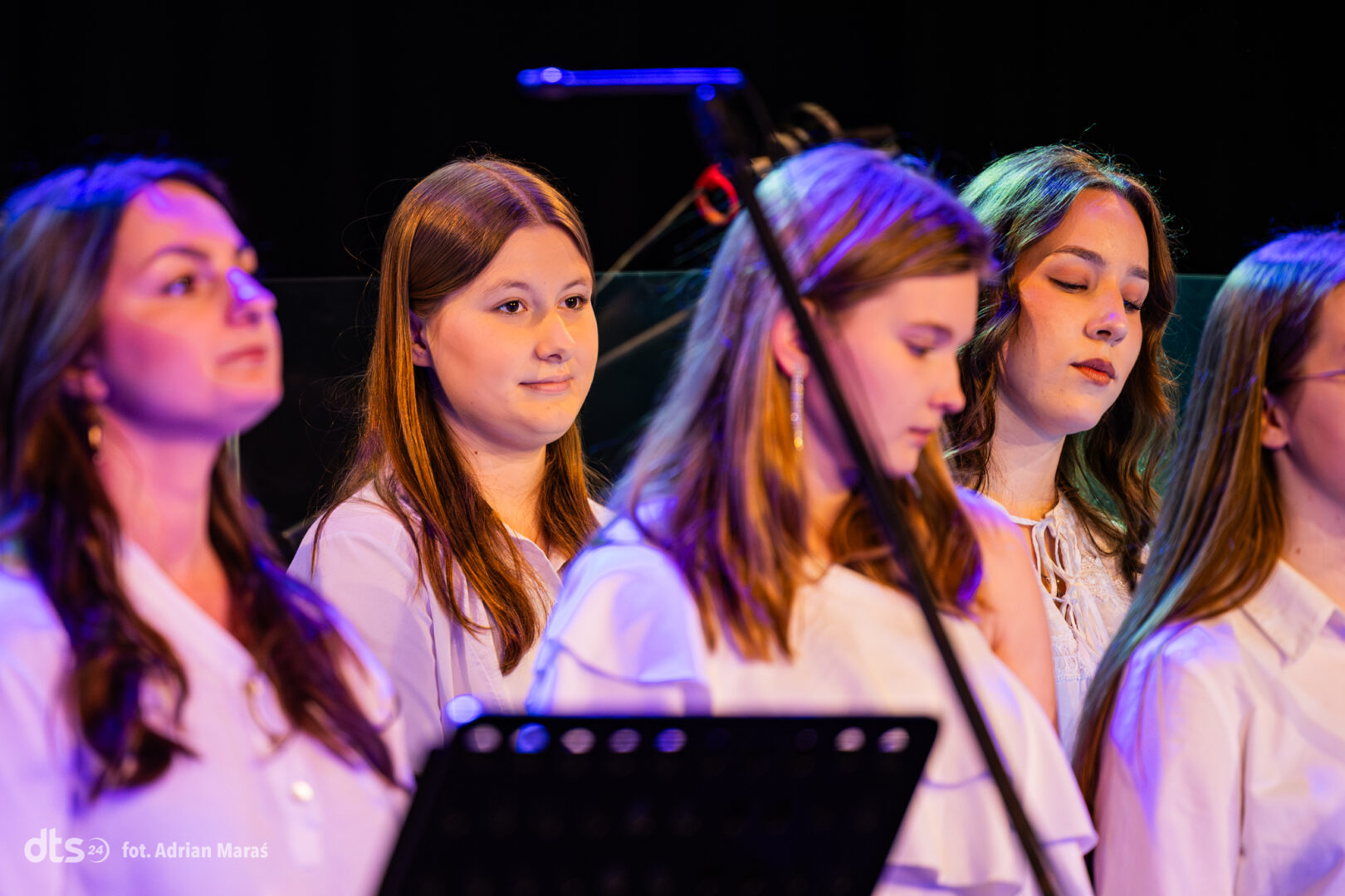 Group of young women in white shirts standing on a dimly lit stage with purple lighting, singing in a choir.