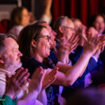 Cheering audience members clapping in a theater with red seats and stage lighting