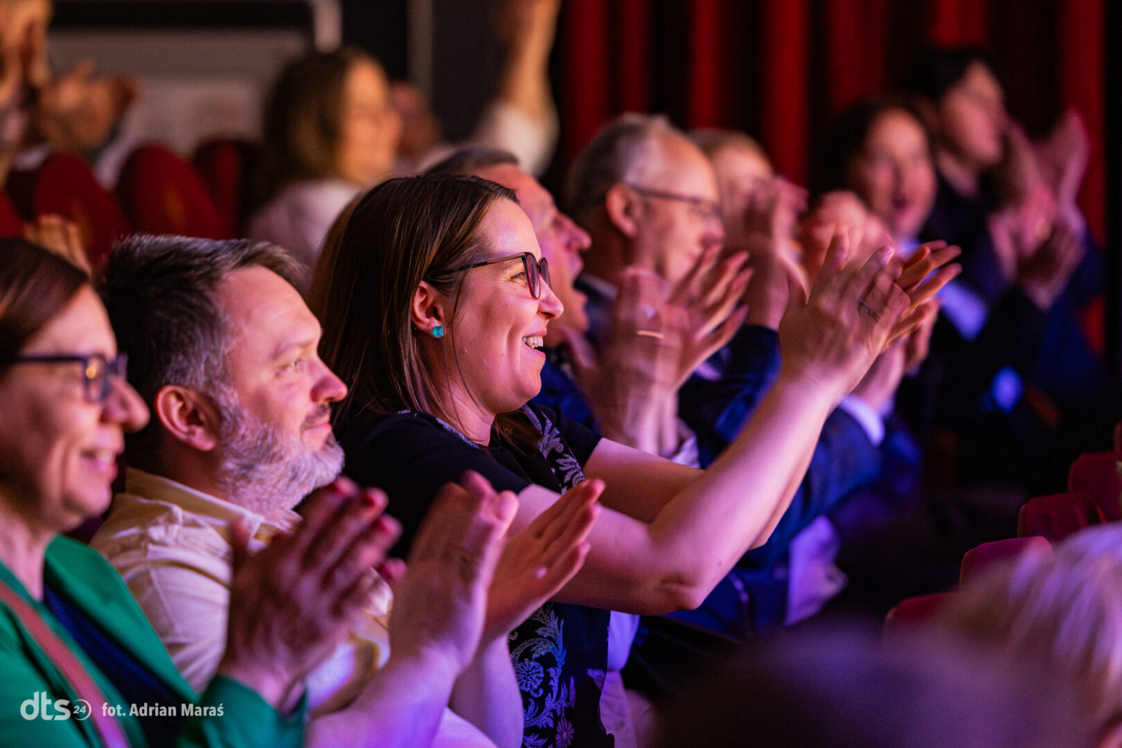 Cheering audience members clapping in a theater with red seats and stage lighting