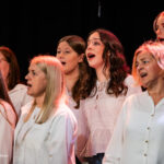 Group of women singing on stage, dressed in white blouses, lit by stage lights.