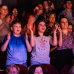 Three children in the front row clapping at a performance: a boy in a blue shirt left, a girl in a light purple shirt center, and a girl in a patterned top right, with an audience behind them.