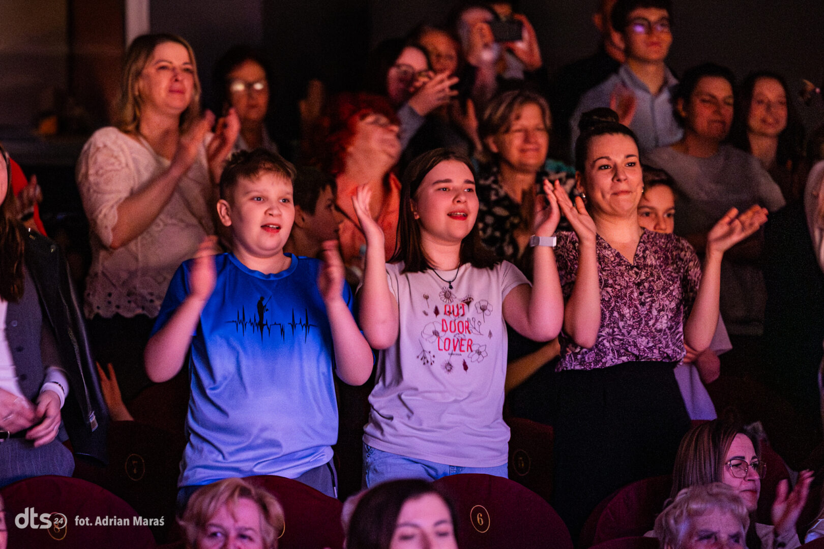 Three children in the front row clapping at a performance: a boy in a blue shirt left, a girl in a light purple shirt center, and a girl in a patterned top right, with an audience behind them.