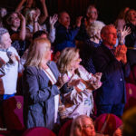 Audience members standing and clapping in a theater during a performance.