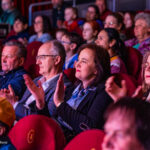 Audience members in a dim theater, seated in red chairs and clapping during a performance.