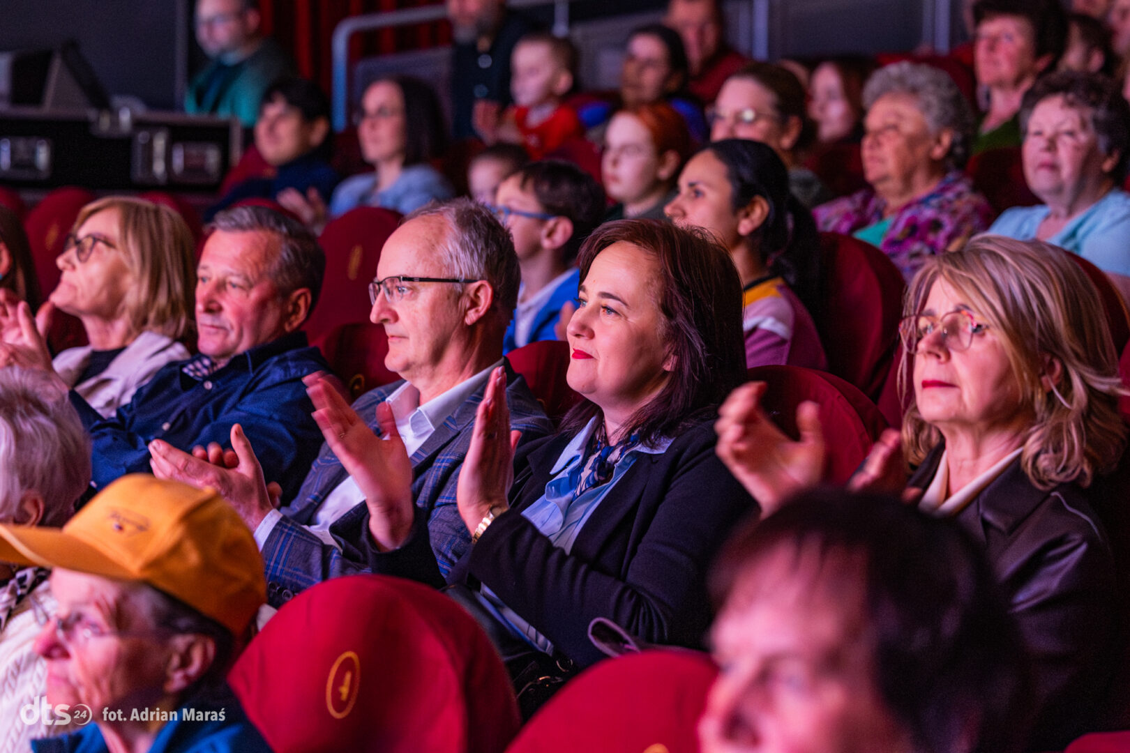 Audience members in a dim theater, seated in red chairs and clapping during a performance.