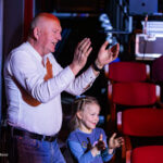 Man and girl clapping together in a theater backstage area, red seats visible in the background, smiling.