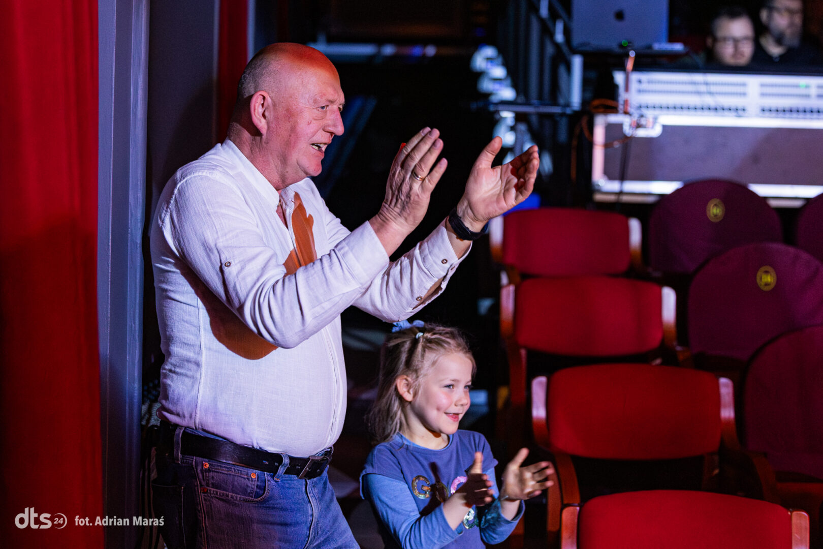 Man and girl clapping together in a theater backstage area, red seats visible in the background, smiling.