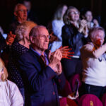 Older man in a blue suit clapping among an audience at a theater.