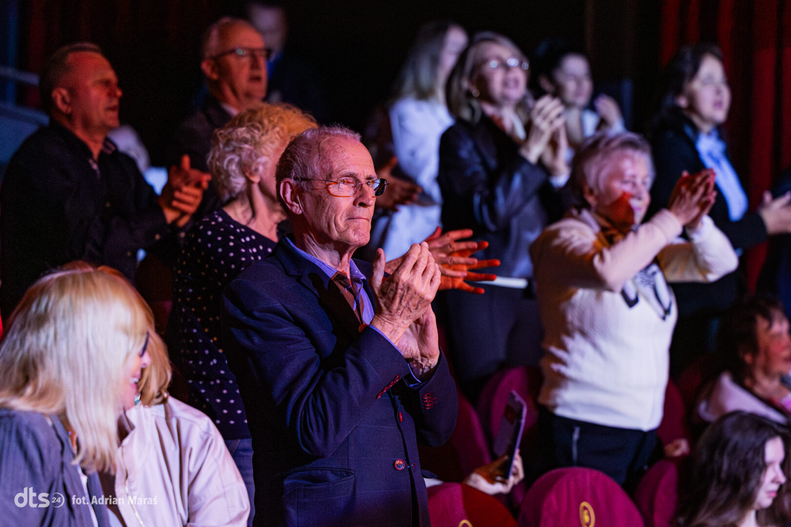 Older man in a blue suit clapping among an audience at a theater.