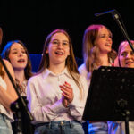 Group of young female singers performing on a dimly lit stage, center-front girl with glasses and white shirt clapping while others sing behind her.