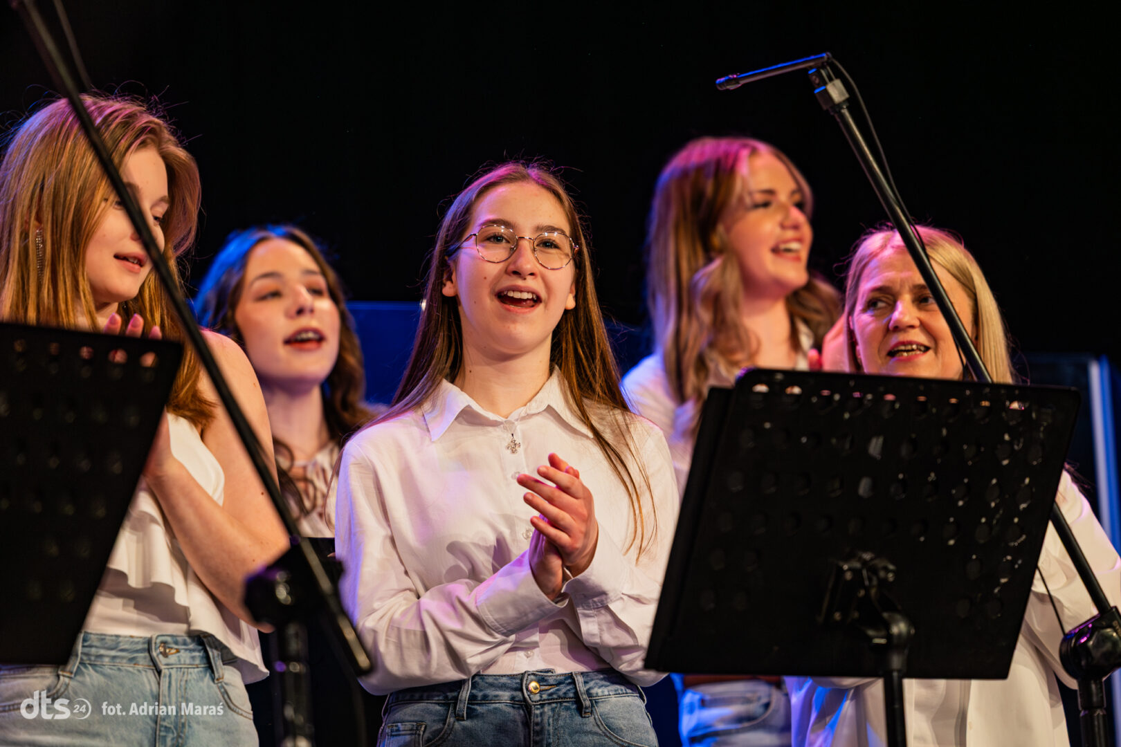 Group of young female singers performing on a dimly lit stage, center-front girl with glasses and white shirt clapping while others sing behind her.