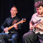 Two male musicians seated on a dim stage, one playing a bass guitar (left) in a black shirt and jeans, the other playing an electric guitar (right) in a patterned shirt.