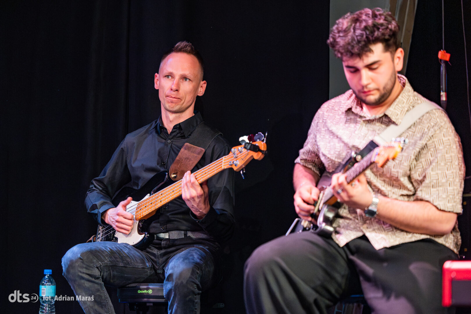 Two male musicians seated on a dim stage, one playing a bass guitar (left) in a black shirt and jeans, the other playing an electric guitar (right) in a patterned shirt.