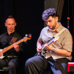 Two male musicians perform on a stage: one bassist on the left and one guitarist on the right, with a red curtain behind them.