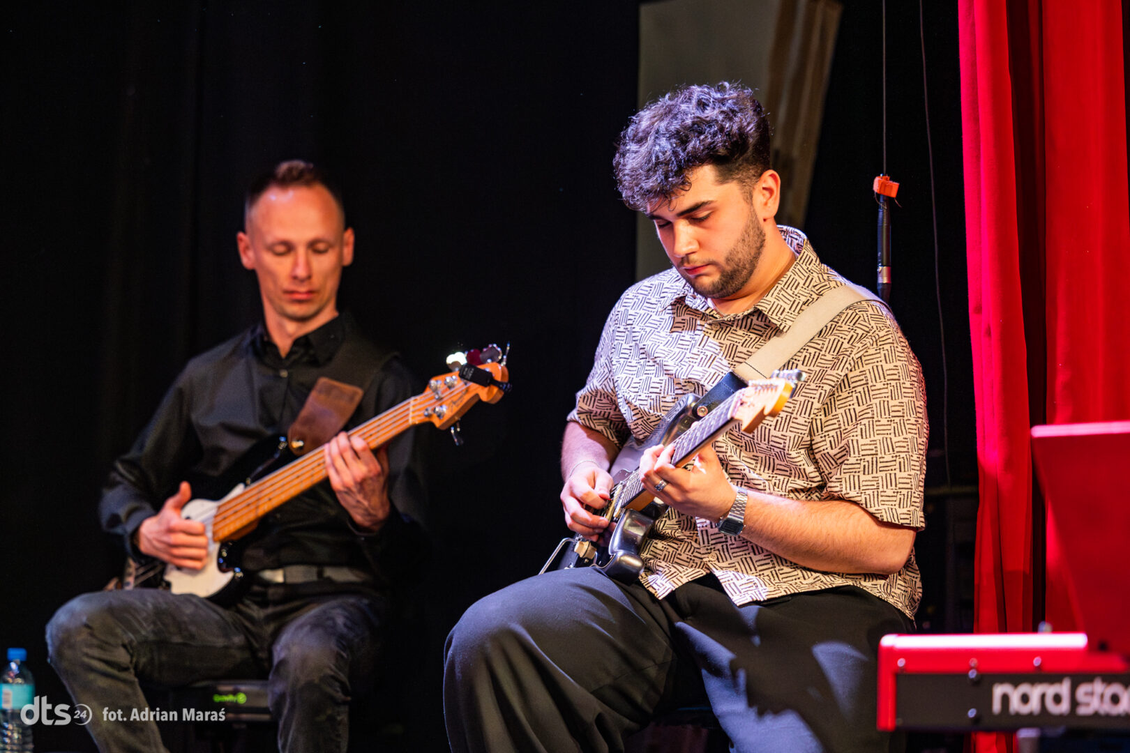 Two male musicians perform on a stage: one bassist on the left and one guitarist on the right, with a red curtain behind them.