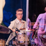 Young drummer wearing glasses plays a drum kit on stage, with sticks mid-air and a clear shield around the kit.