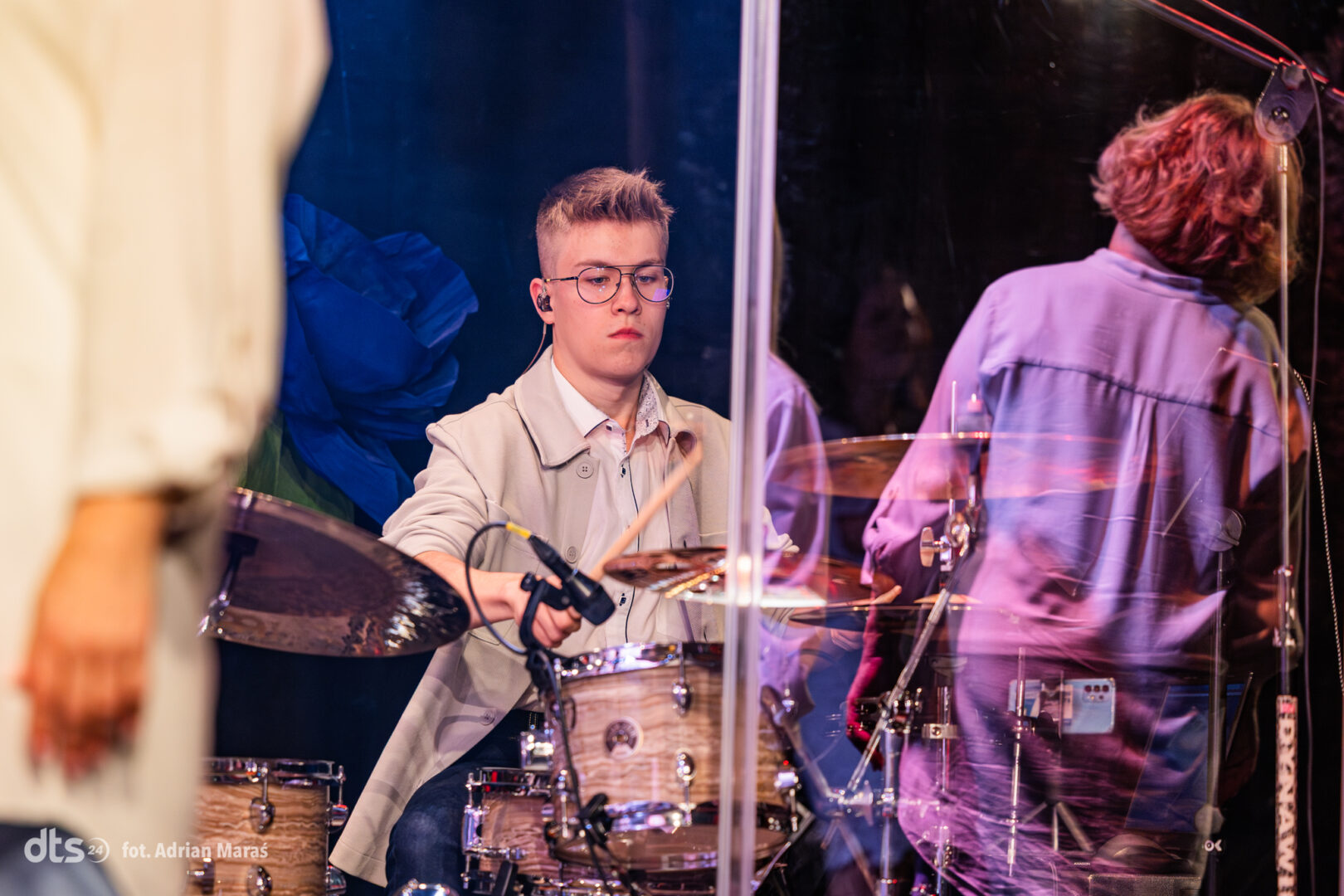 Young drummer wearing glasses plays a drum kit on stage, with sticks mid-air and a clear shield around the kit.
