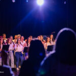 A choir of children and adults singing on a stage under a blue spotlight, audience silhouettes in the foreground.