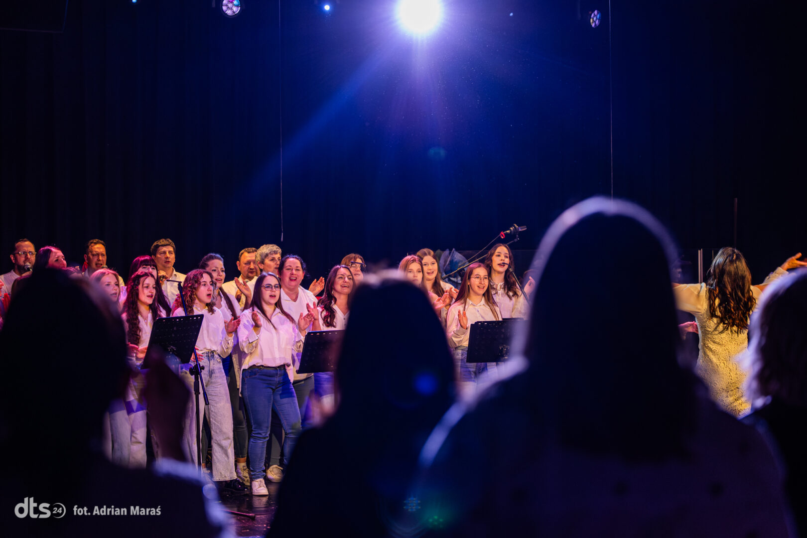 A choir of children and adults singing on a stage under a blue spotlight, audience silhouettes in the foreground.
