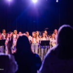 Choir of children singing onstage under blue-purple lights, facing microphones with music stands, audience silhouettes in foreground.