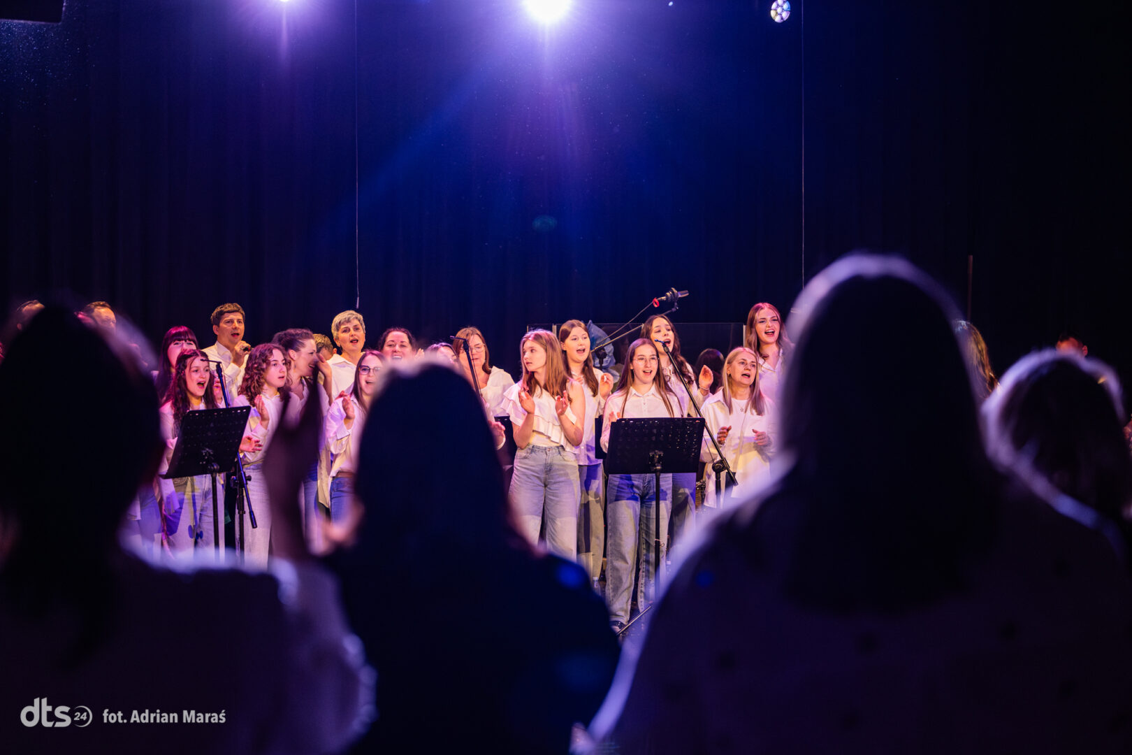 Choir of children singing onstage under blue-purple lights, facing microphones with music stands, audience silhouettes in foreground.