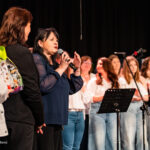 Woman in a blue blouse speaks into a microphone on stage, with a choir behind her.