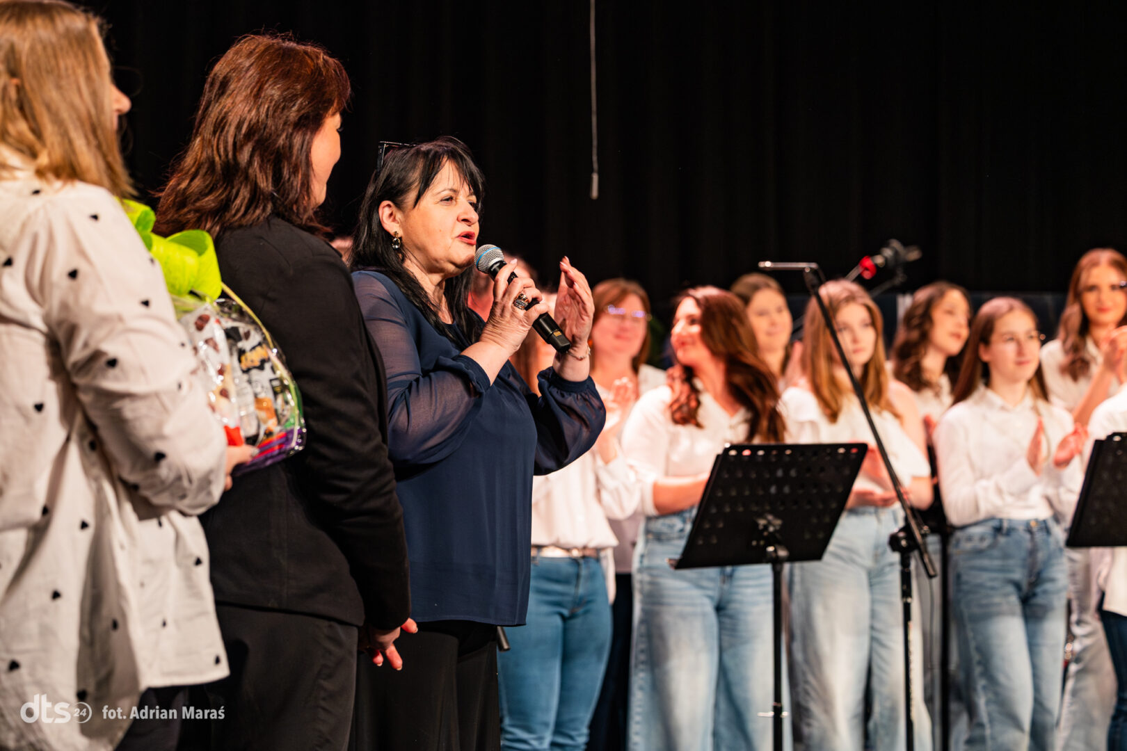 Woman in a blue blouse speaks into a microphone on stage, with a choir behind her.
