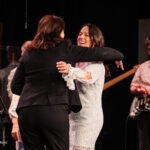 Two people embrace and dance on a stage, smiling, with a guitarist watching in the background