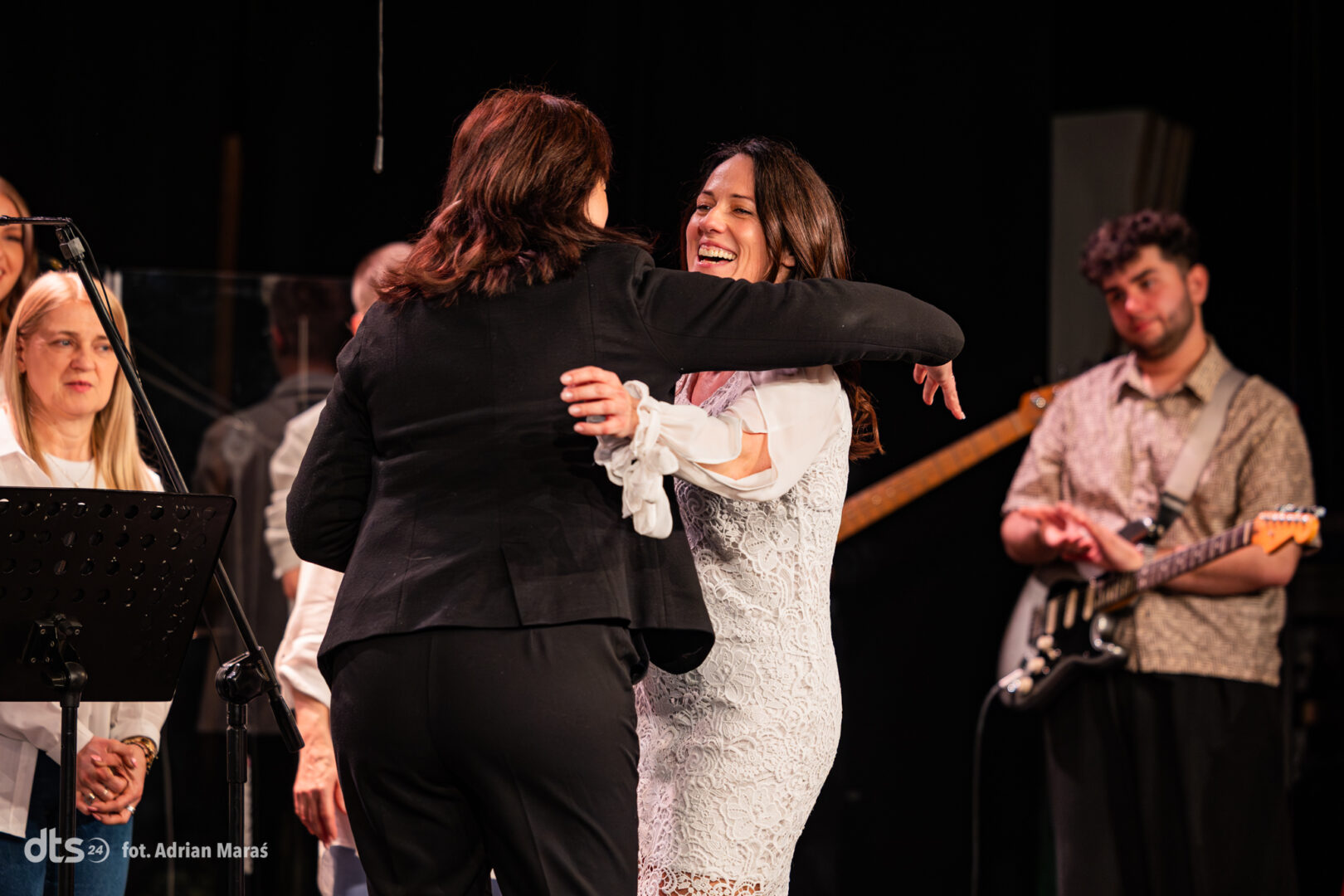 Two people embrace and dance on a stage, smiling, with a guitarist watching in the background