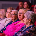 Audience of seniors watching a performance in a dim theater; foreground woman in bright pink top wearing glasses looks focused on the stage.