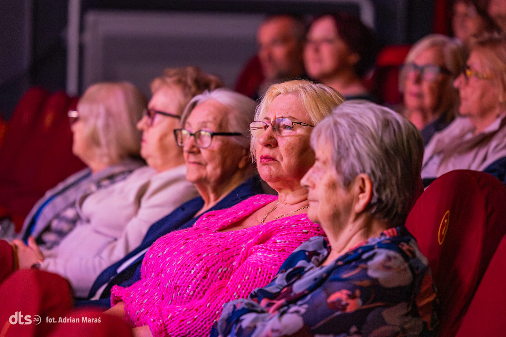Audience of seniors watching a performance in a dim theater; foreground woman in bright pink top wearing glasses looks focused on the stage.
