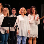 Group of performers on stage: a smiling woman in a white lace dress claps while others stand with microphones and music stands nearby, a guitarist on the right.