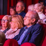Audience of seniors watching a performance in a red-seat theater.