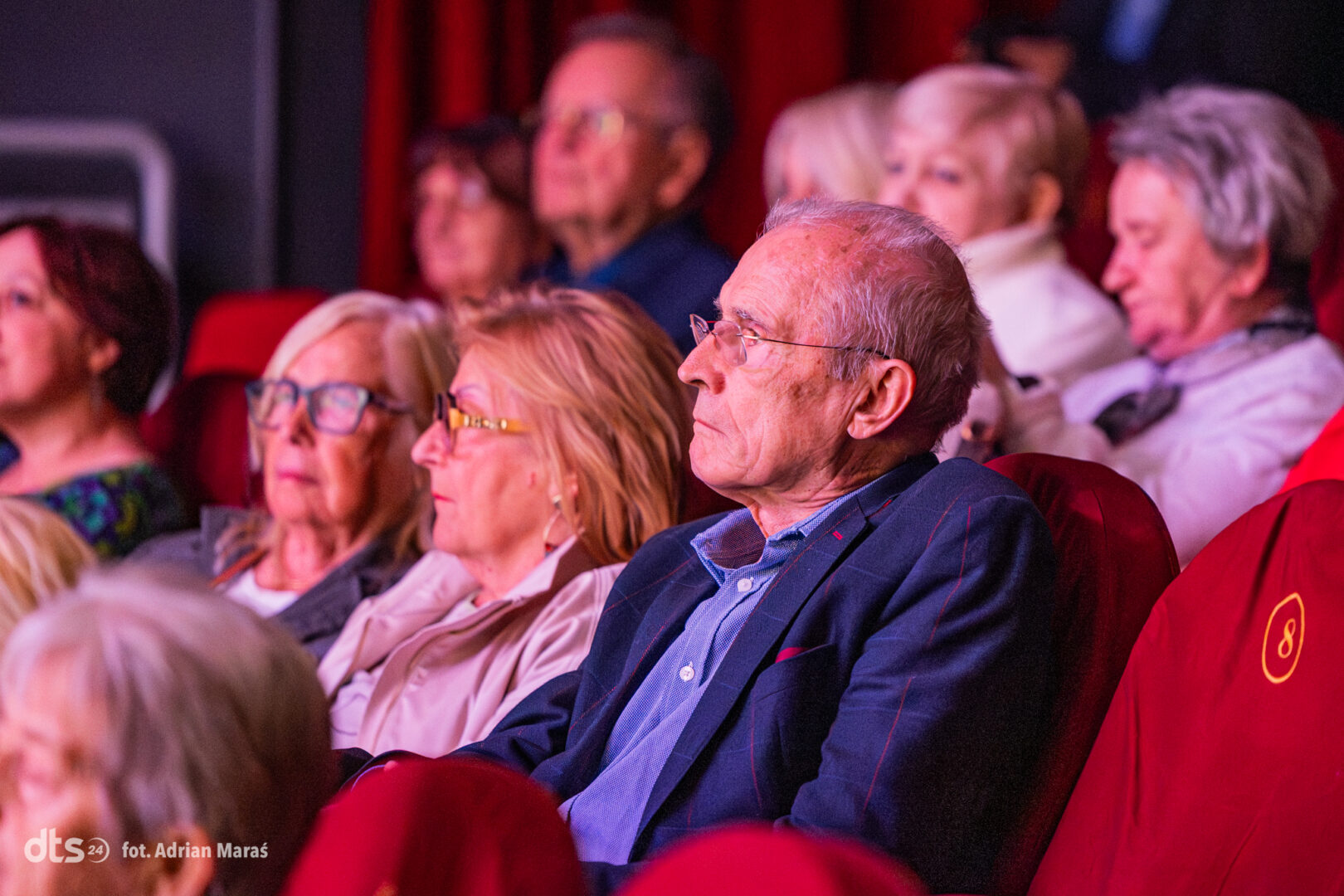 Audience of seniors watching a performance in a red-seat theater.