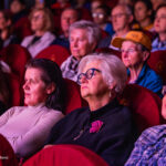 Audience seated in a dimly lit theater, faces focused on the stage above them.