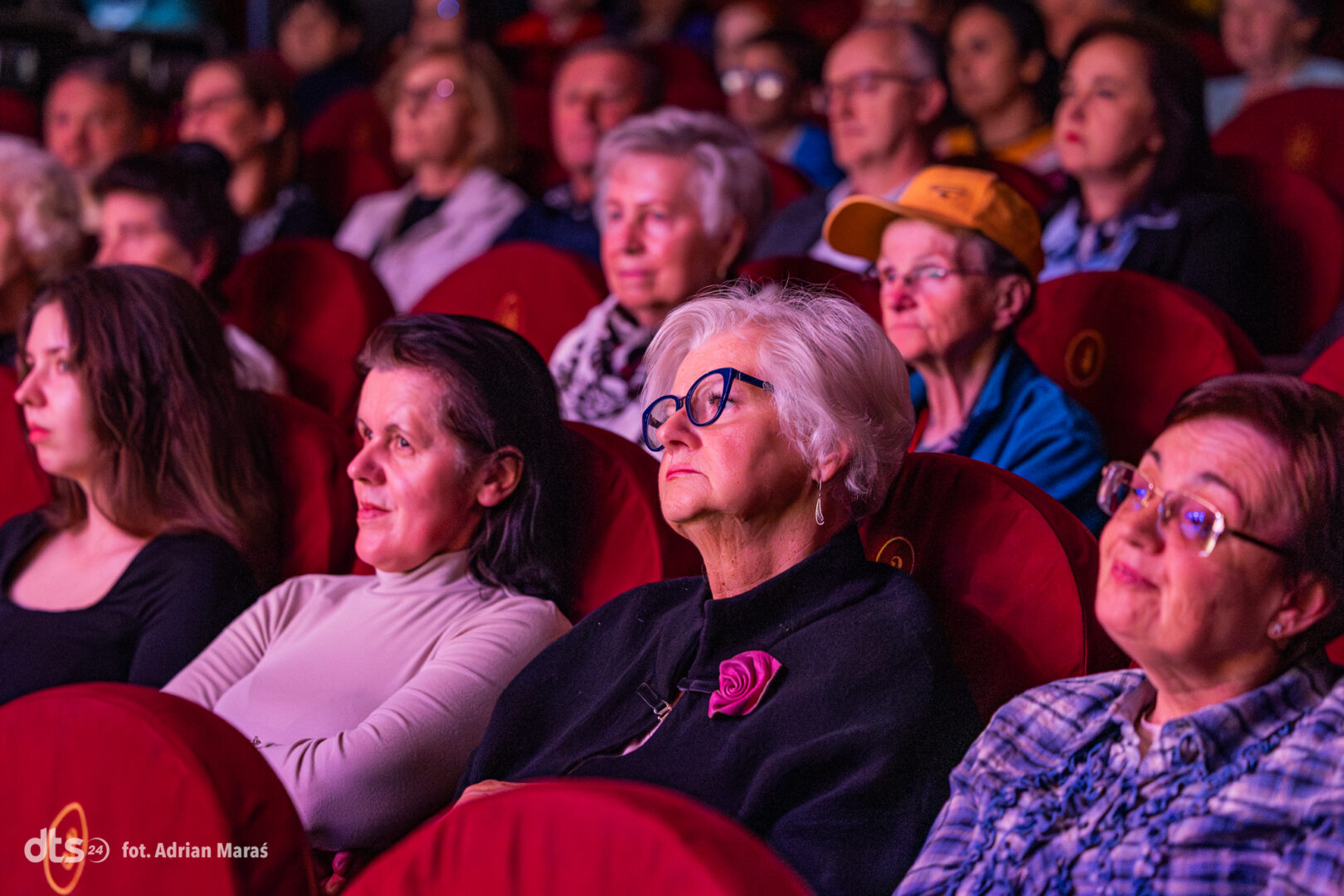 Audience seated in a dimly lit theater, faces focused on the stage above them.