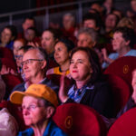 Audience members in red theater seats watching a show and clapping.
