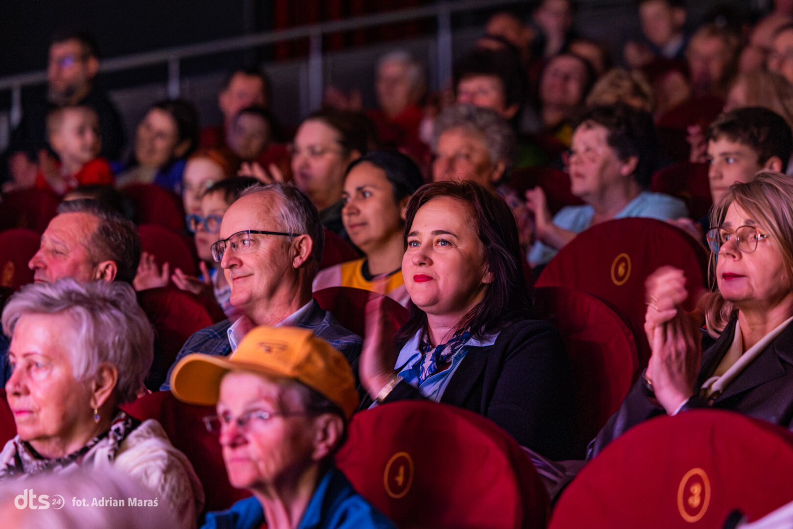 Audience members in red theater seats watching a show and clapping.