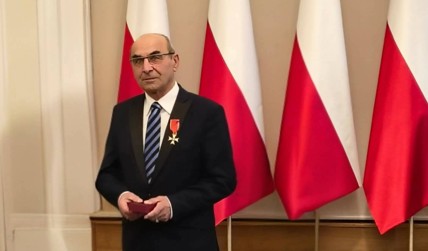 Older man in a dark suit with striped tie stands at a podium, wearing a medal on his chest and holding a small red case, with Polish flags in the background.