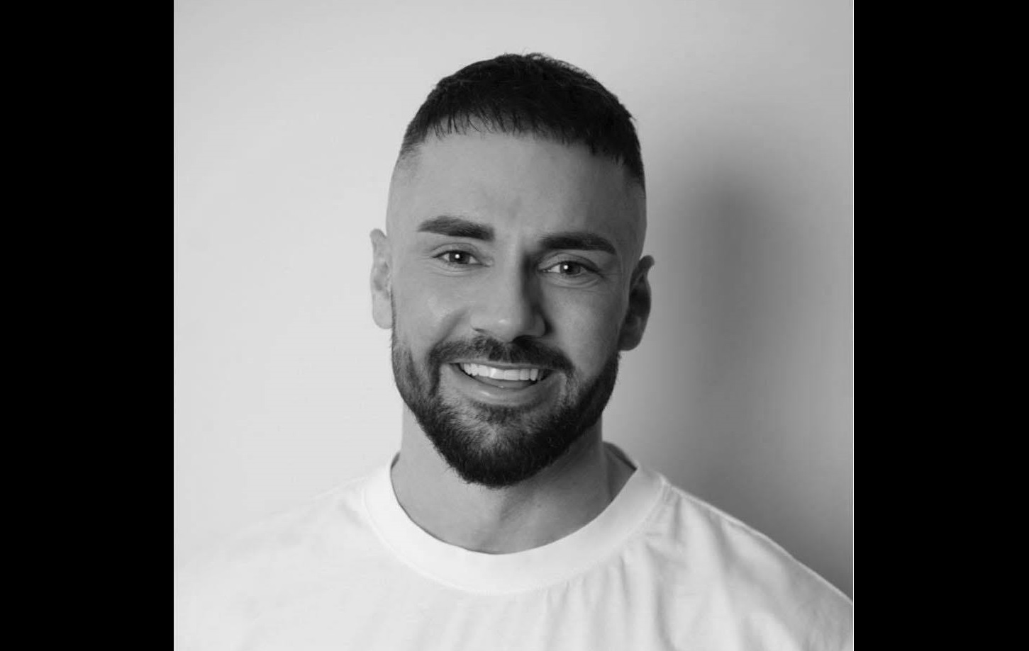 Smiling man with a trimmed beard and short hair in a white T-shirt, portrait against a light background (black-and-white).