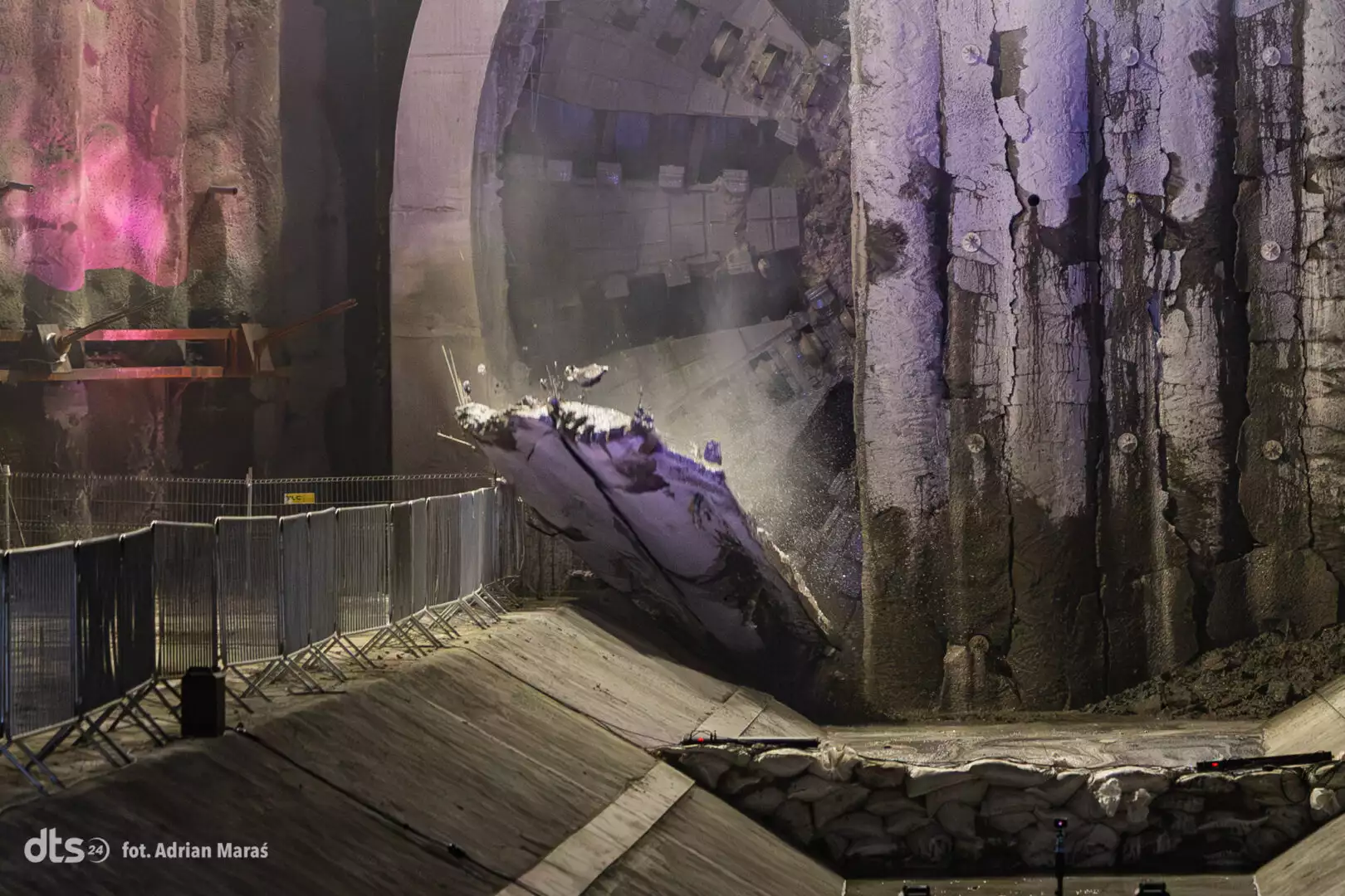 Night view of a massive quarry wall with a broken concrete slab near a circular tunnel entrance and safety barriers along a sloped road at the ground level.