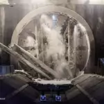 Tunnel boring machine in a rock excavation, circular TBM shield with water spray and flying debris inside the tunnel chamber, safety barriers along the sides.