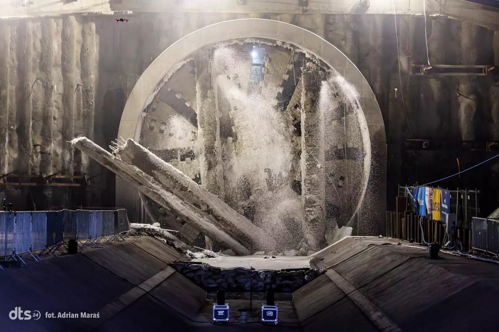 Tunnel boring machine in a rock excavation, circular TBM shield with water spray and flying debris inside the tunnel chamber, safety barriers along the sides.